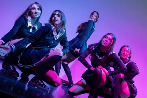 Cinco jóvenes con uniforme escolar de falda y americana posan agachadas sobre un escenario iluminado con luz morada y rosa.