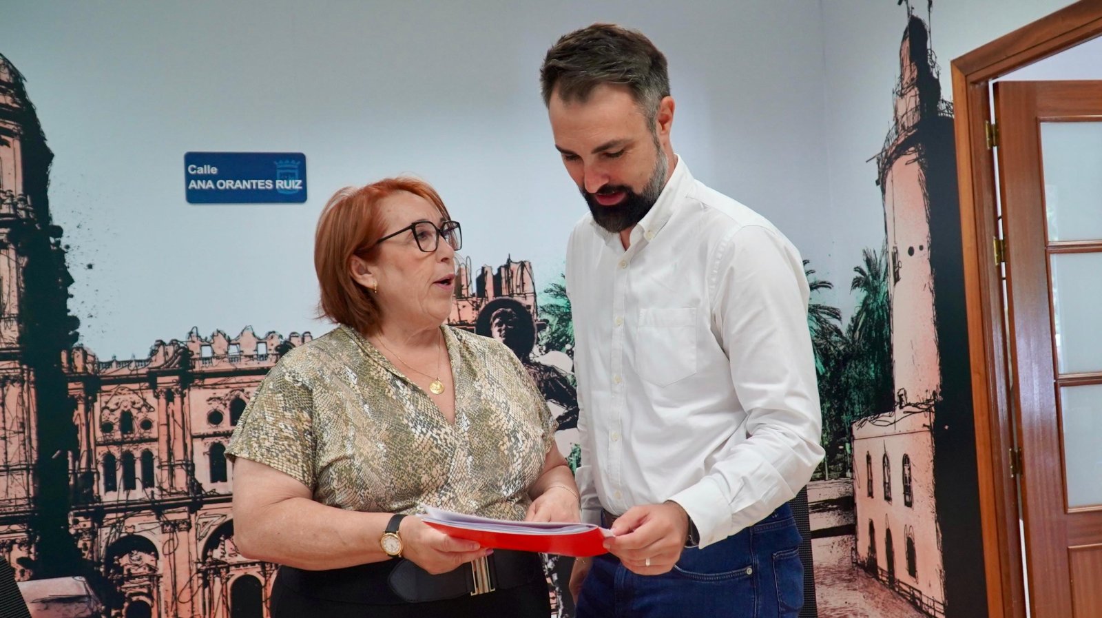 Una mujer con gafas y un hombre con camisa blanca sostienen una carpeta roja ante un mural con edificios y una placa de calle.