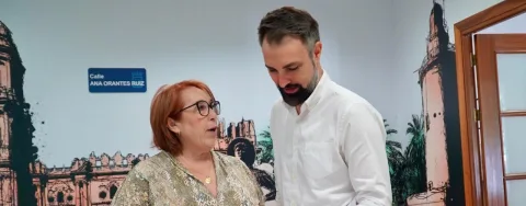 Una mujer mayor con gafas y un hombre joven con barba, de pie ante una pared con un cartel de la calle Ana Orantes Ruiz y un dibujo de edificios.