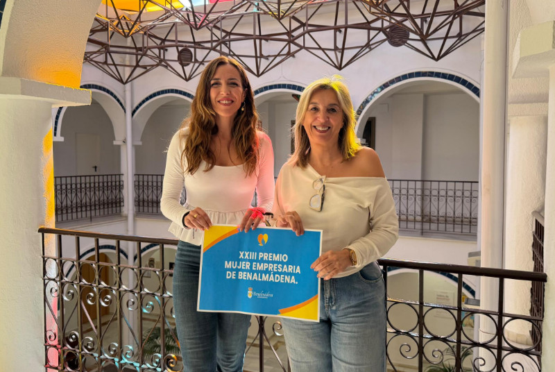 Dos mujeres sonrientes sostienen un cartel del XXIII Premio Mujer Empresaria de Benalmádena en un patio con arcos.