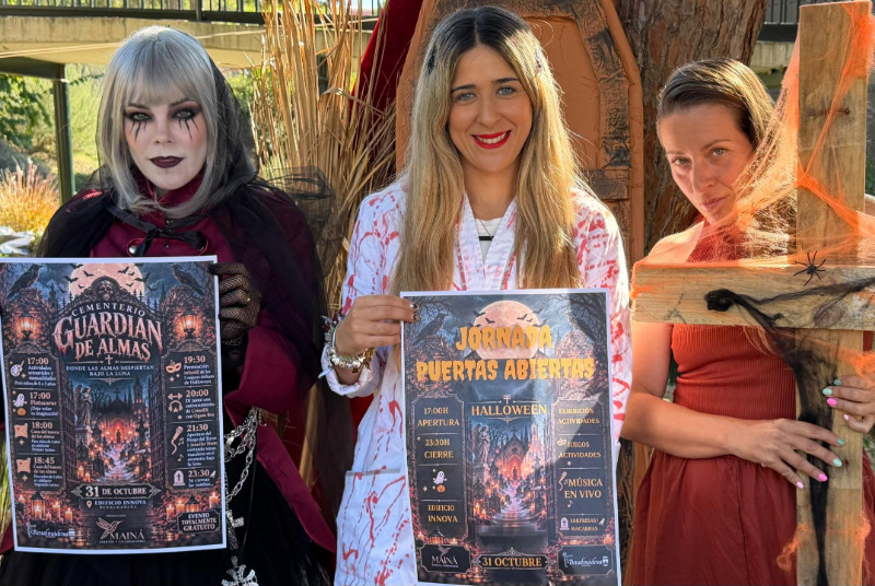 Tres mujeres disfrazadas de Halloween sostienen carteles de un evento y una cruz de madera con telarañas.