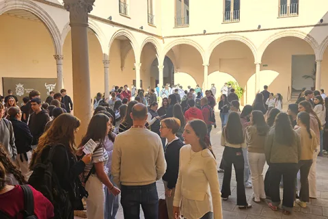 Una multitud de jóvenes de pie conversando en un patio porticado con arcos y columnas de piedra.