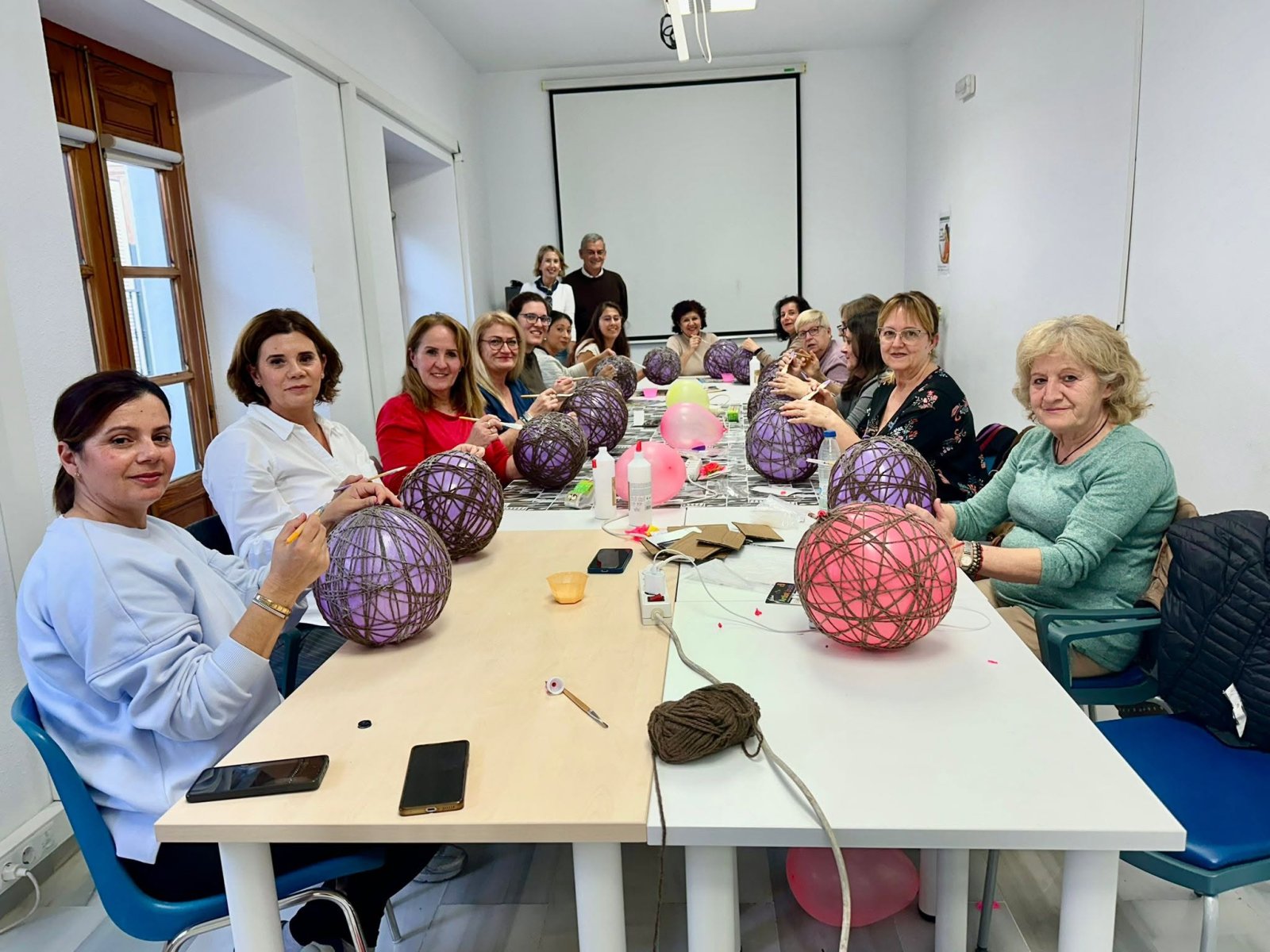 Un grupo de mujeres sentadas alrededor de una mesa larga en un aula, manipulando esferas de hilo morado y rosa sobre globos.