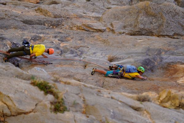 Dos escaladores con casco y arnés ascienden por una pared de roca, uno tumbado y otro de pie, unidos por una cuerda.