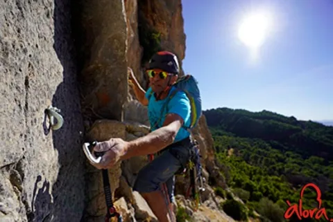 Un escalador con casco, gafas y mochila asciende por una pared rocosa equipada con cintas, con un valle boscoso y sol al fondo.