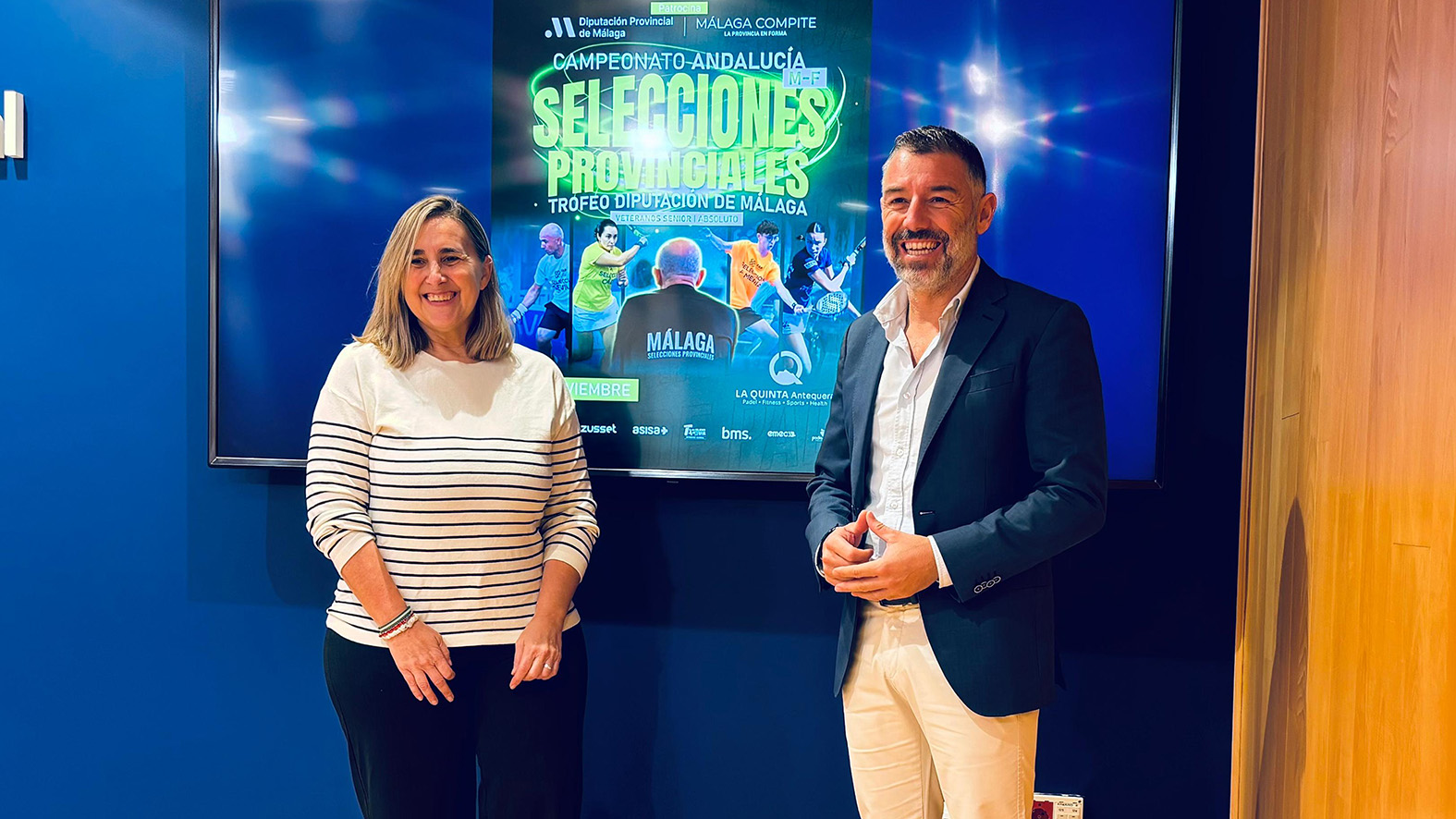 Un hombre y una mujer de pie, sonriendo, delante de una pantalla con un cartel del Campeonato de Andalucía de Selecciones Provinciales.