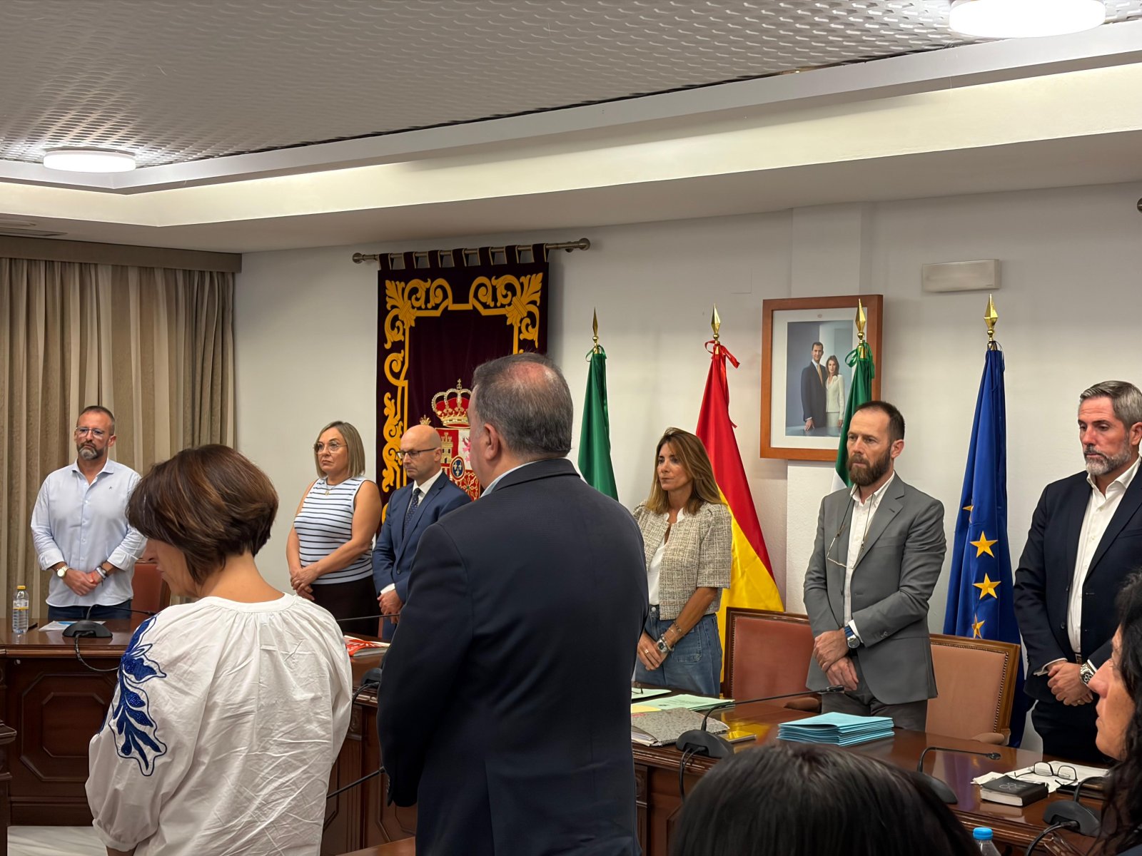 Varias personas de pie en una sala con banderas de España, Andalucía y la UE, un retrato enmarcado y una mesa de reuniones.