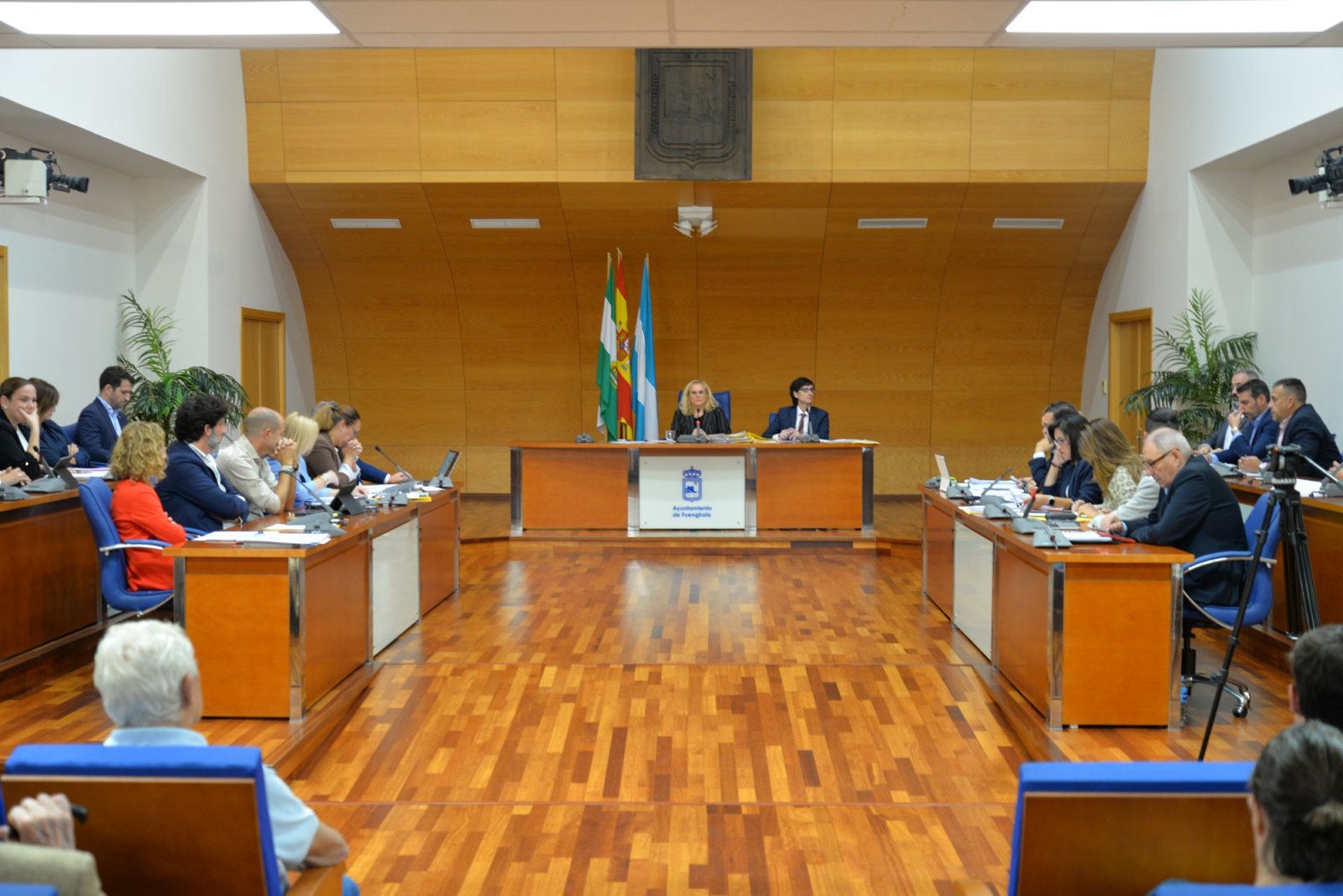 Sala de plenos con varias personas sentadas en mesas enfrentadas y dos presidiendo al fondo junto a banderas.