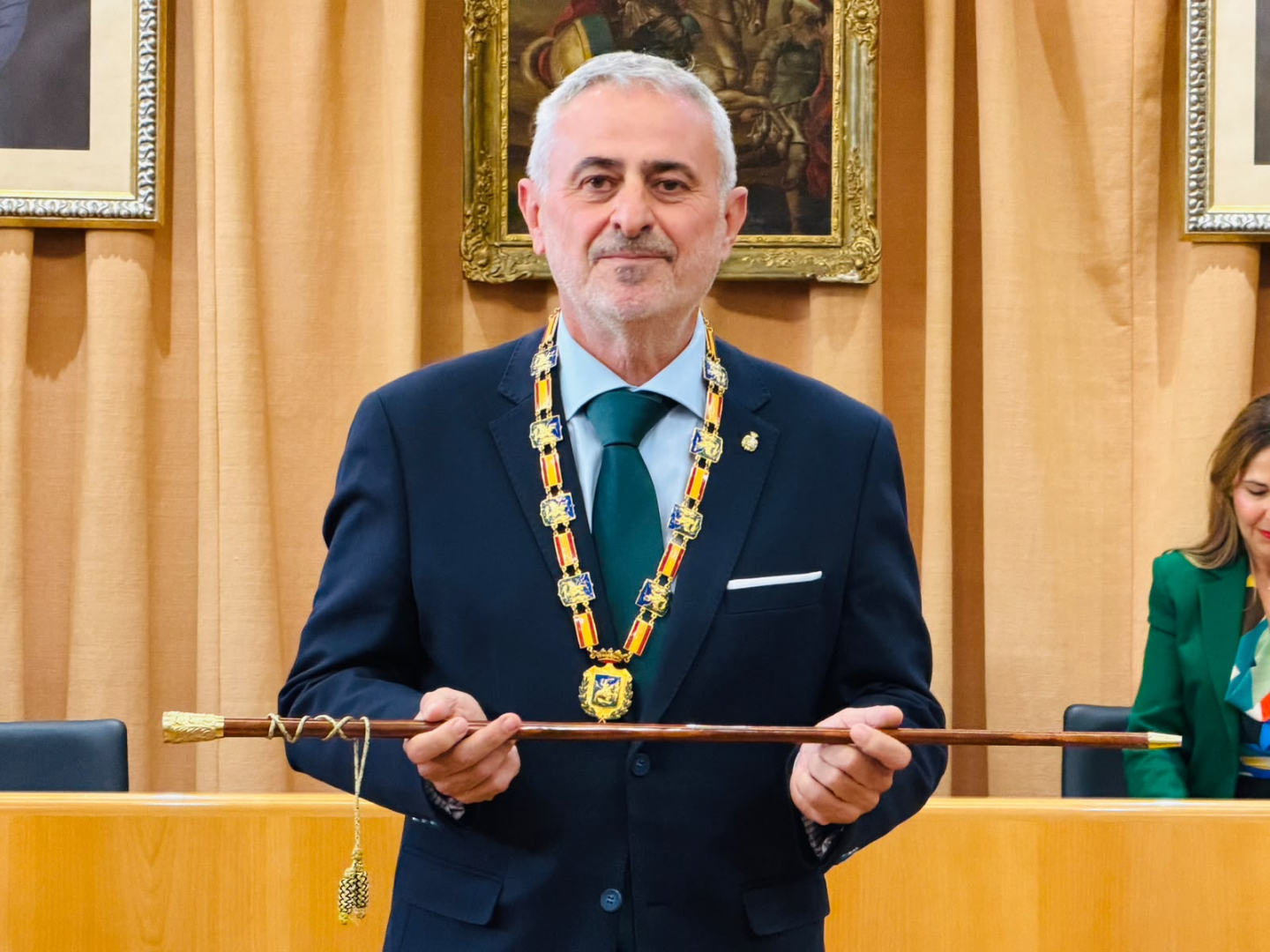 Hombre de traje azul con corbata verde y collar ceremonial sostiene un bastón de mando ante una cortina y cuadros.