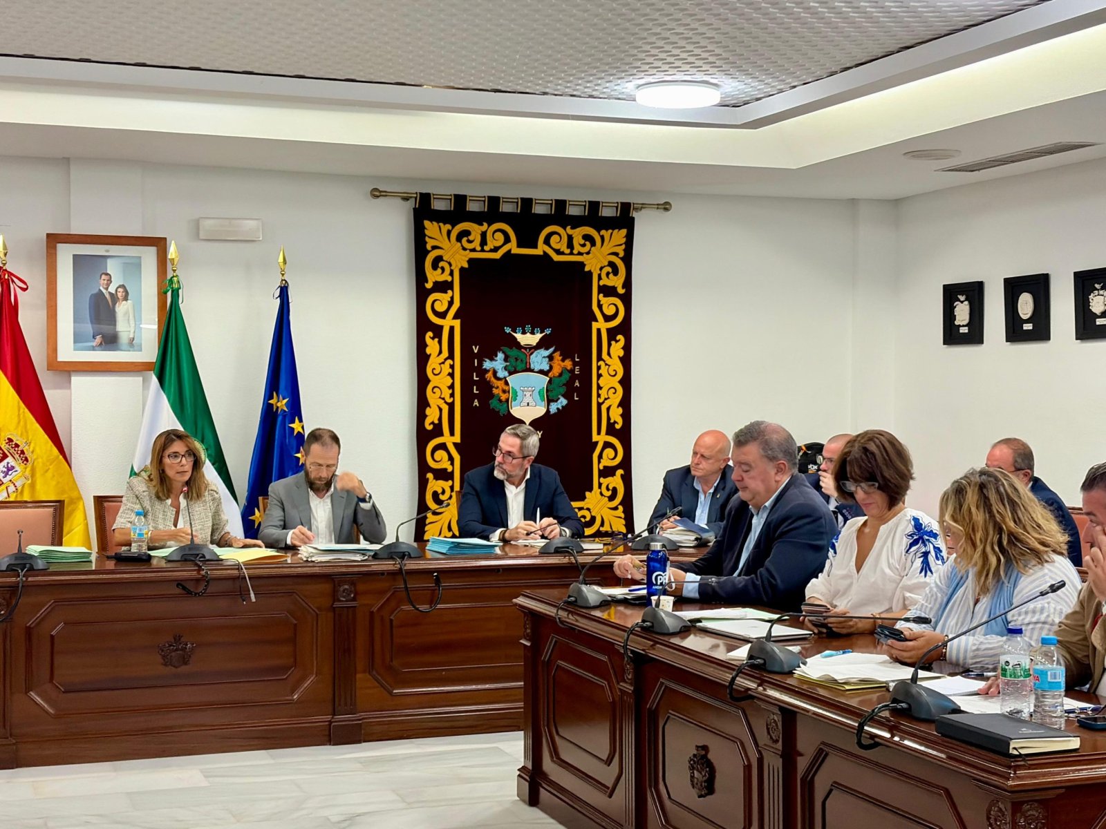 Varias personas sentadas tras una mesa de madera en una sala con banderas de España, Andalucía y la UE y un escudo bordado al fondo.