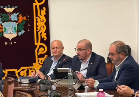 Tres hombres trajeados sentados ante micrófonos y tablets en una mesa de reunión, con un escudo heráldico bordado al fondo.