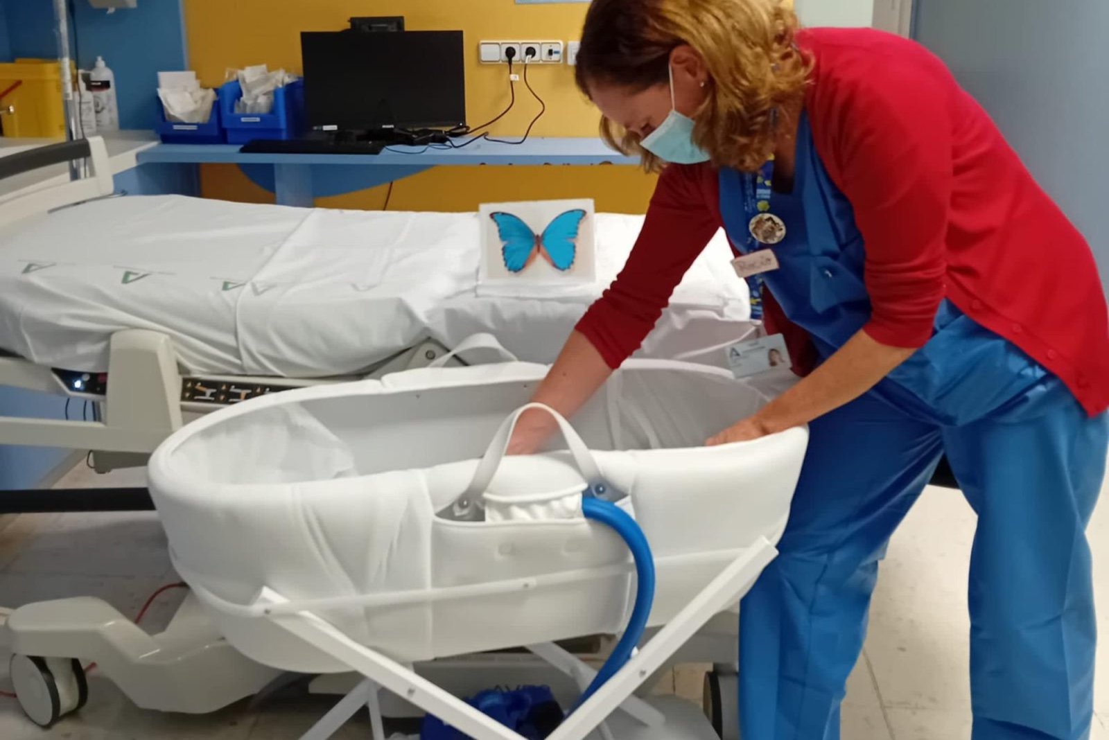Una sanitaria con mascarilla y pijama azul se inclina junto a una cuna hospitalaria blanca, en una habitación con una cama y una mariposa azul.