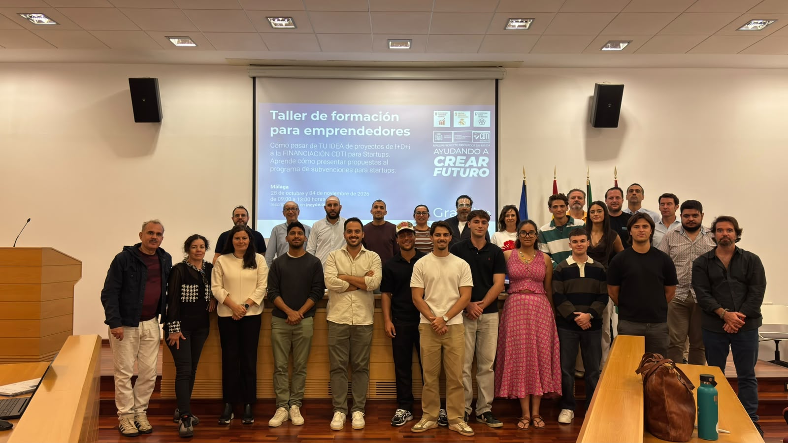 Un grupo de personas posa de pie en una sala con una pantalla de proyección al fondo que anuncia un taller de formación para emprendedores.