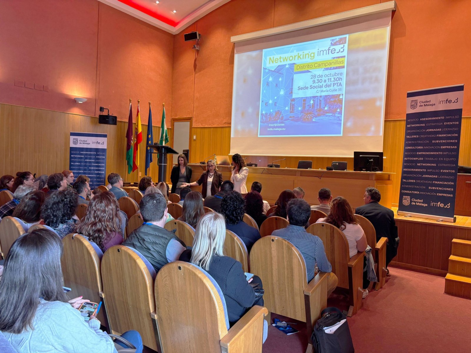 Público sentado en un salón de actos frente a una pantalla que proyecta una sesión de networking y tres personas de pie junto a la mesa.