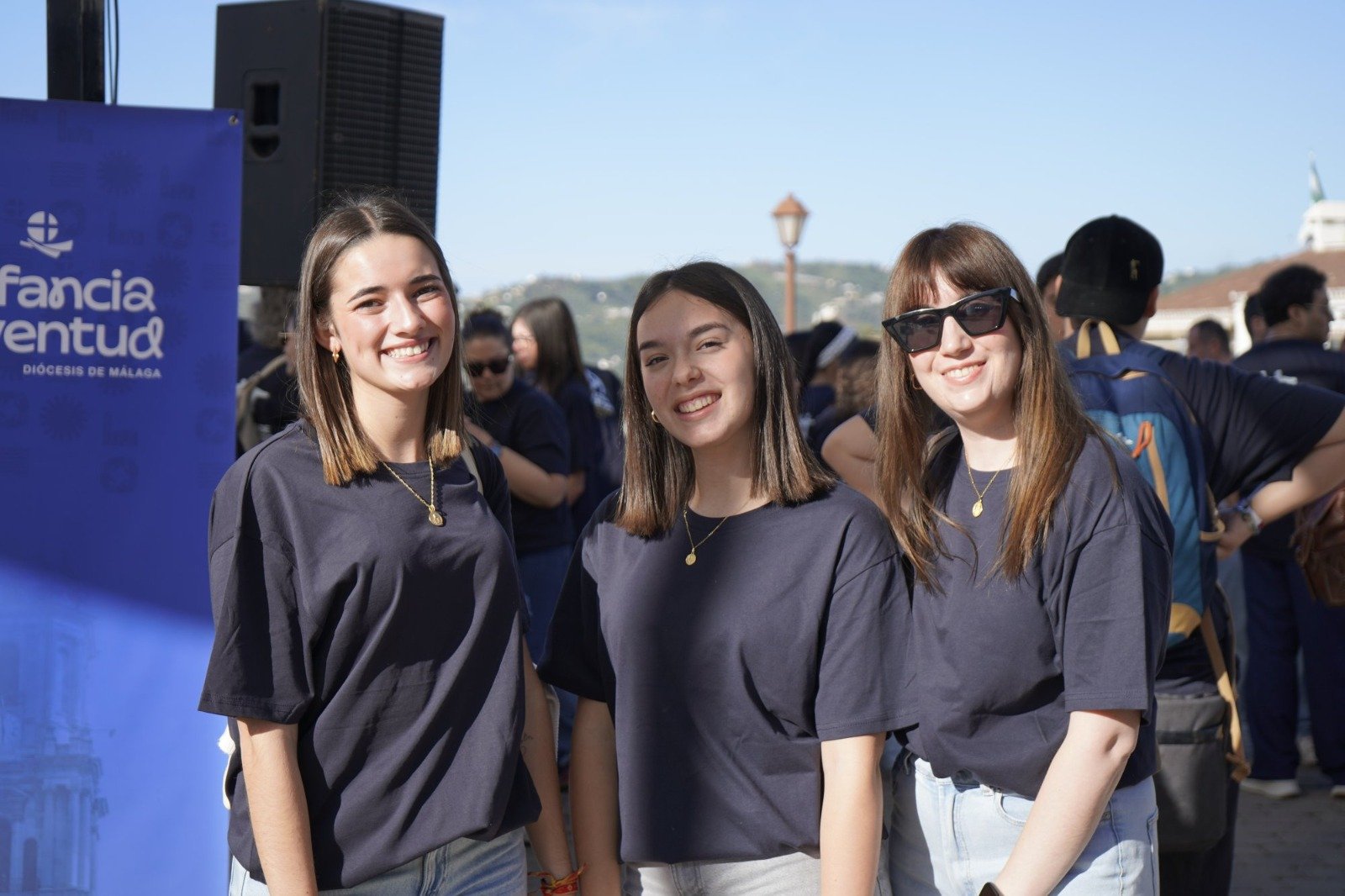 Tres jóvenes con camiseta negra sonríen al aire libre junto a un cartel azul y un altavoz, con más gente al fondo.