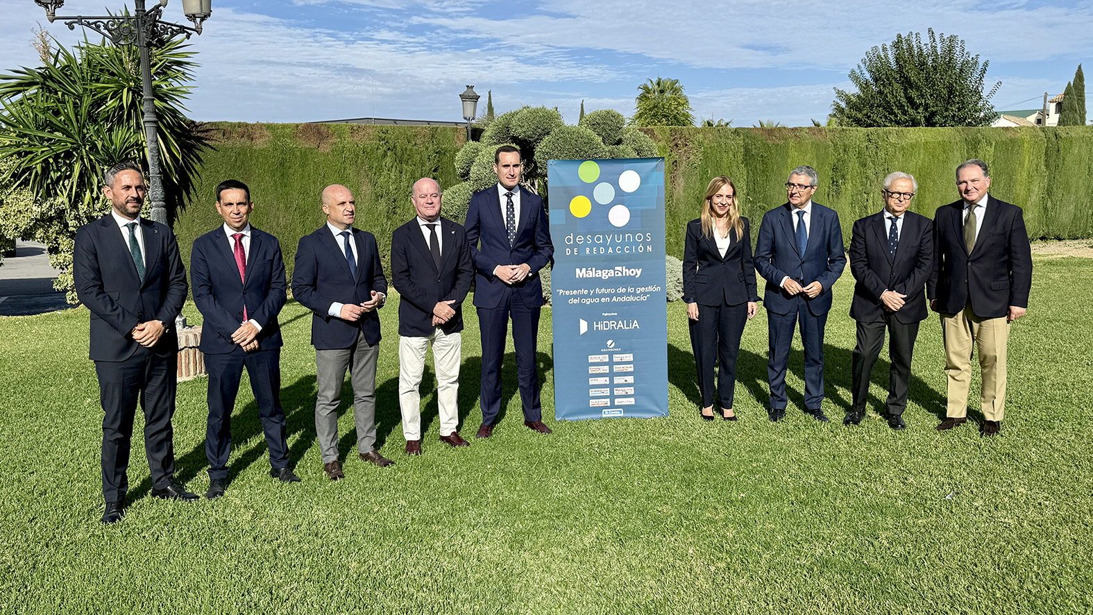 Nueve personas trajeadas posan en un jardín junto a un cartel vertical sobre gestión del agua en Andalucía.
