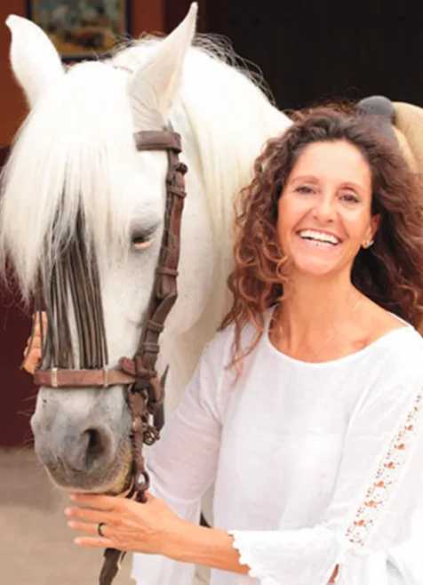 Una mujer sonriente de pelo rizado y blusa blanca sostiene la cabeza de un caballo blanco con cabezada.