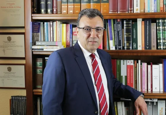 Un hombre con gafas, traje azul y corbata roja posa de pie ante una estantería llena de libros.