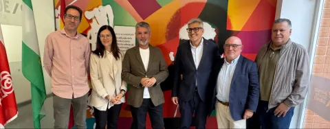 Seis personas de pie, posando en fila ante un mural de colores vivos y una bandera de Andalucía.