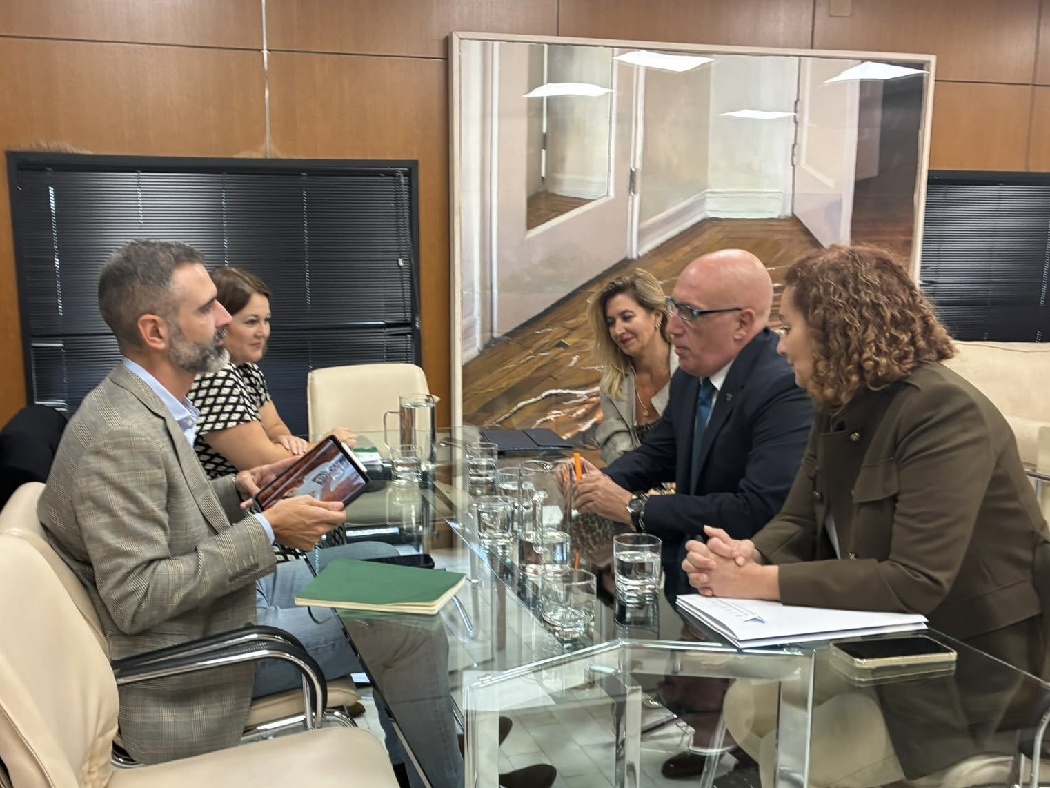 Cuatro personas sentadas alrededor de una mesa de cristal en una sala de reuniones con paredes de madera y un gran cuadro al fondo.