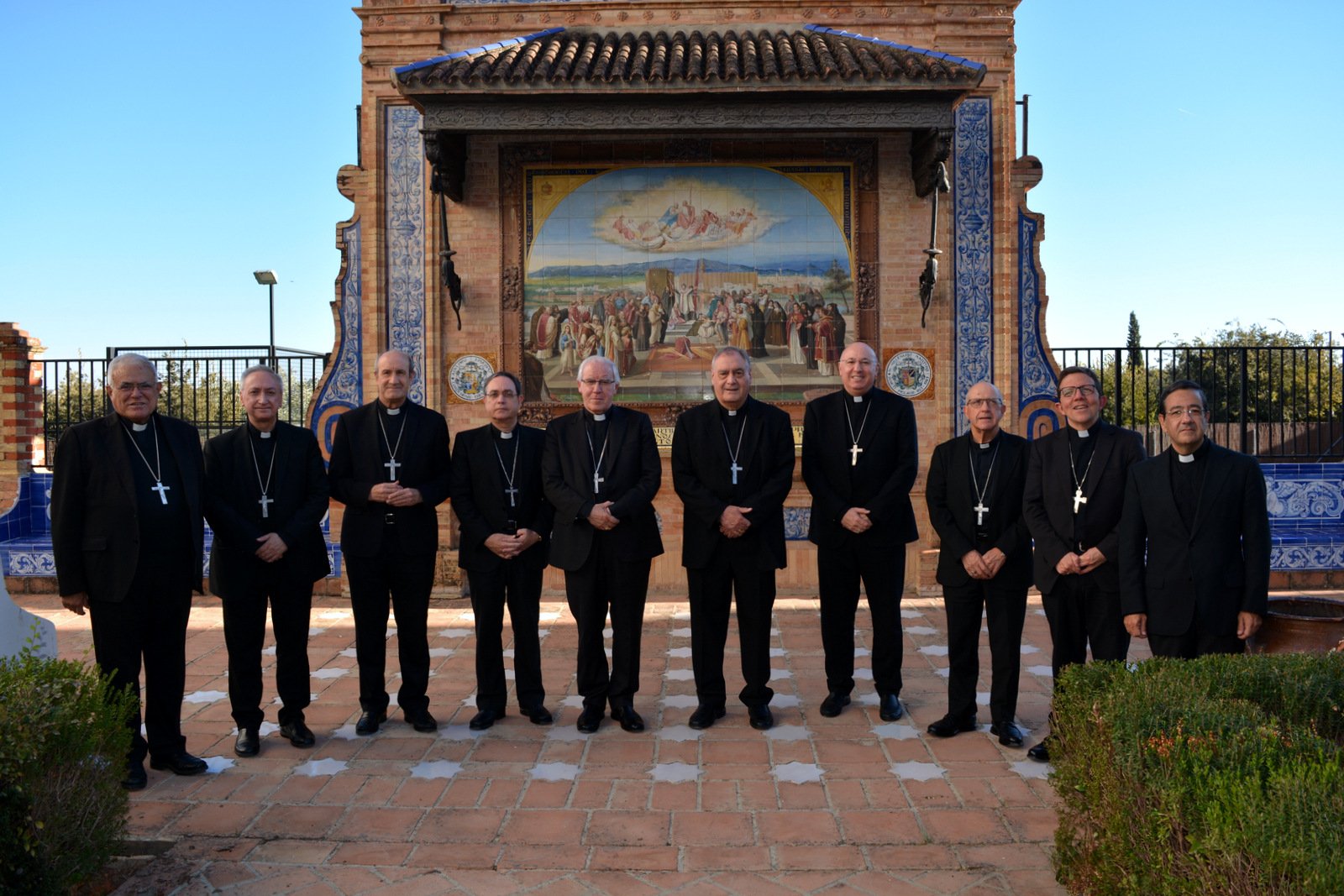 Diez hombres con traje negro y cruz pectoral posan en fila al aire libre ante un retablo cerámico de azulejos con cubierta.