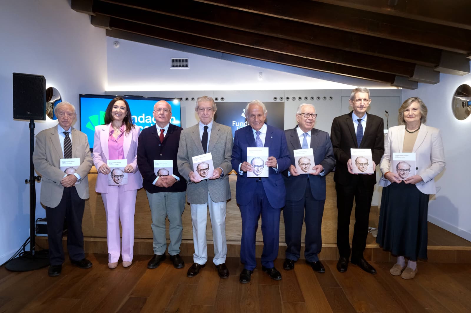 Ocho personas de pie en fila sostienen cada una un libro con el retrato de un hombre en la portada, en una sala con pantalla al fondo.
