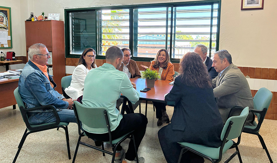 Siete personas sentadas alrededor de una mesa en una sala con ventanas de persianas verdes, hablando entre ellas.