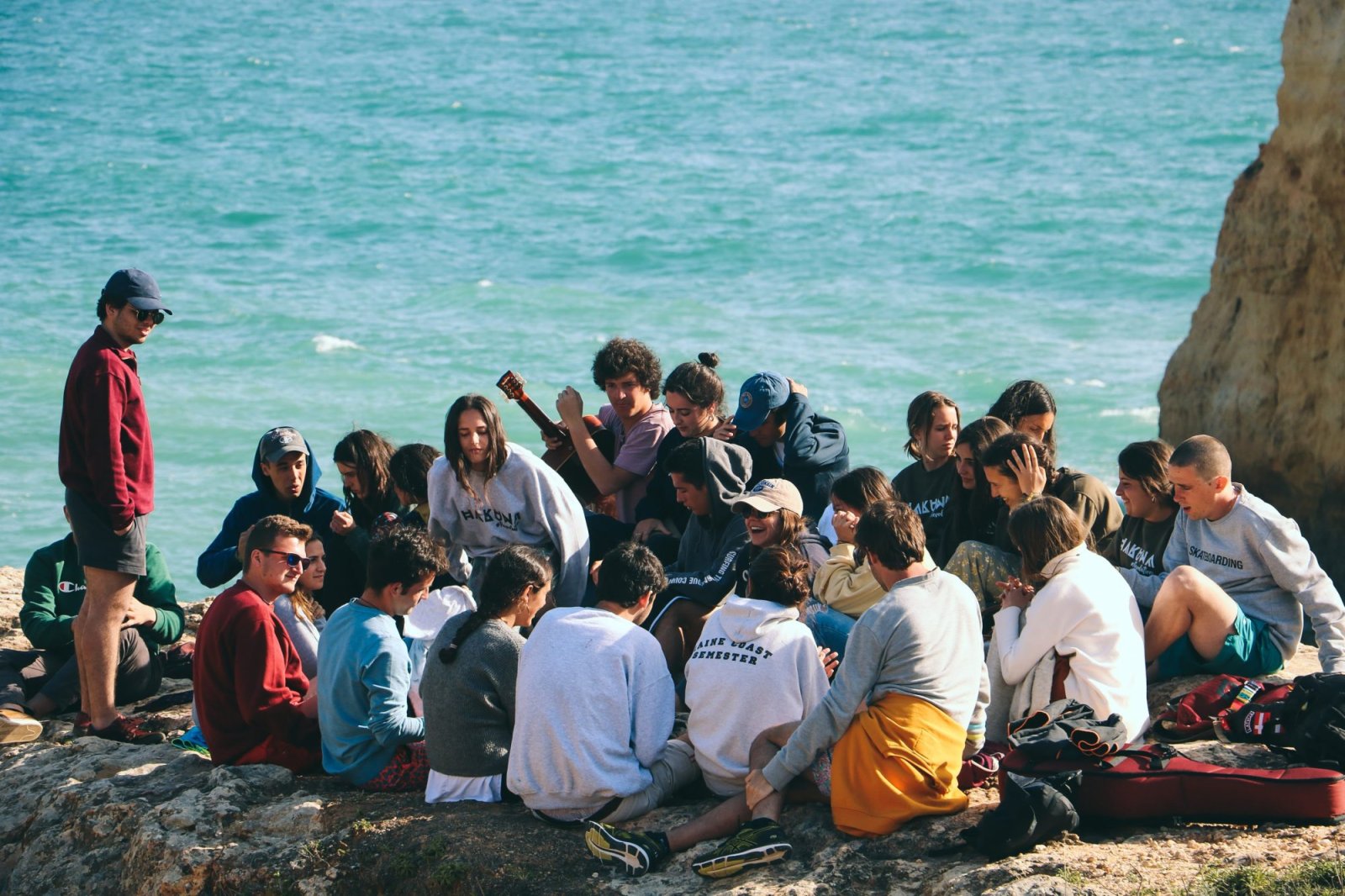 Un grupo de jóvenes sentados en unas rocas junto al mar, algunos con guitarras, junto a un acantilado.
