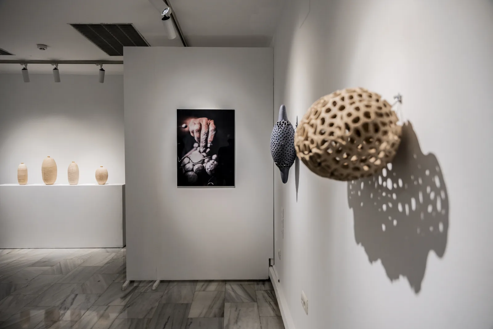 Sala de exposiciones con vasijas sobre una repisa, una fotografía enmarcada en un panel y una pieza esférica calada en la pared.