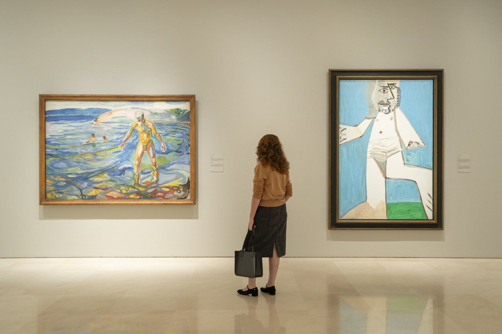 Una mujer de espaldas con bolso negro contempla dos cuadros colgados en la pared blanca de una sala de museo.
