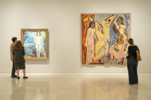 Dos personas contemplan dos cuadros en una sala de museo: uno pequeño de tonos azules y otro grande con figuras femeninas.