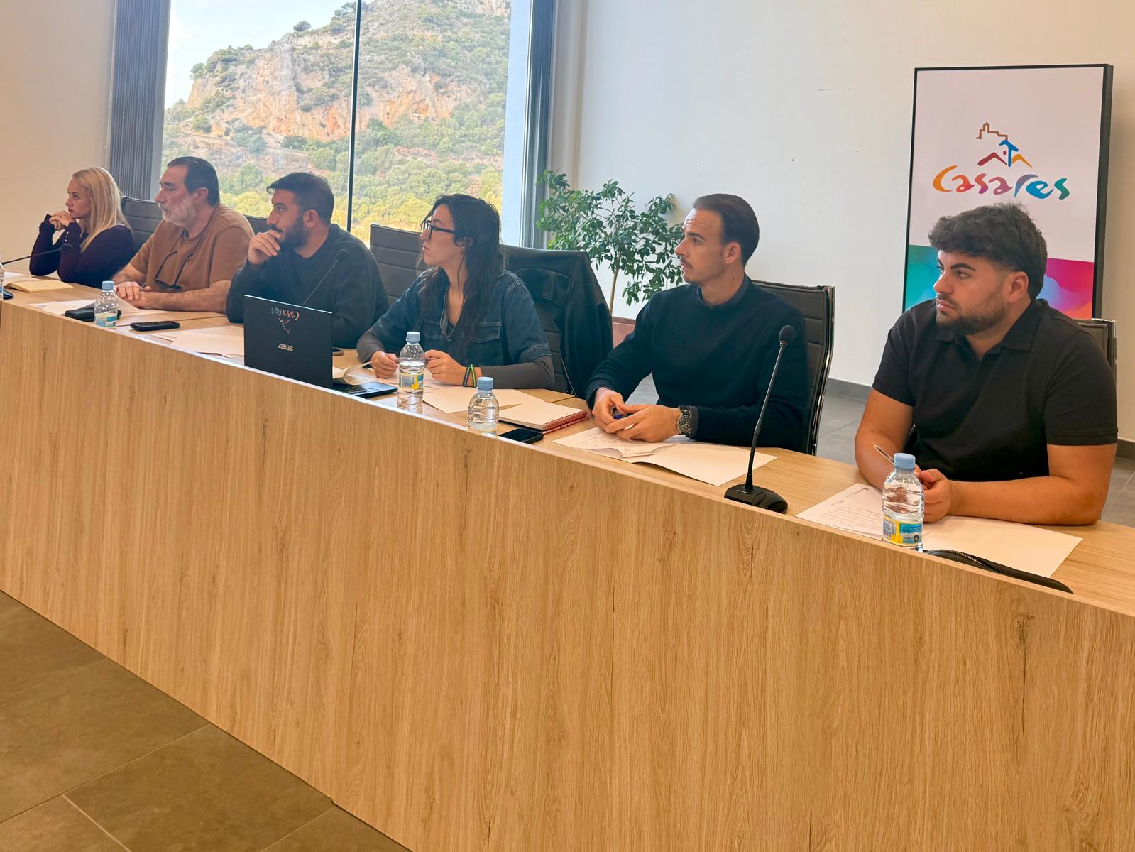 Seis personas sentadas tras una mesa de madera en una sala con ventanal, un cartel con el logotipo de Casares y una planta.