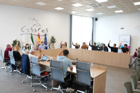 Varias personas sentadas en una mesa de reuniones larga levantan la mano para votar en una sala con banderas y el rótulo Casares.