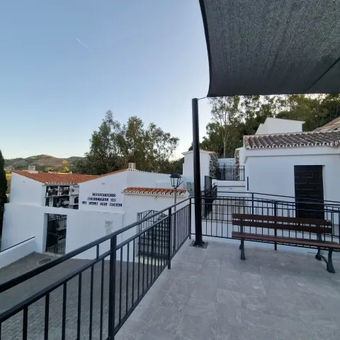 Terraza con barandilla y banco de madera junto a edificios blancos de tejados rojos y árboles al fondo.