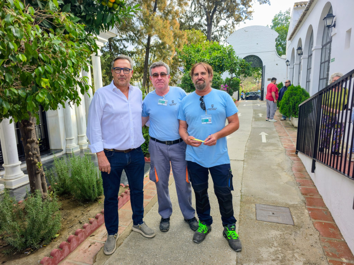 Tres hombres de pie en un camino de cementerio, dos con camiseta azul y acreditación, junto a edificios blancos y árboles.