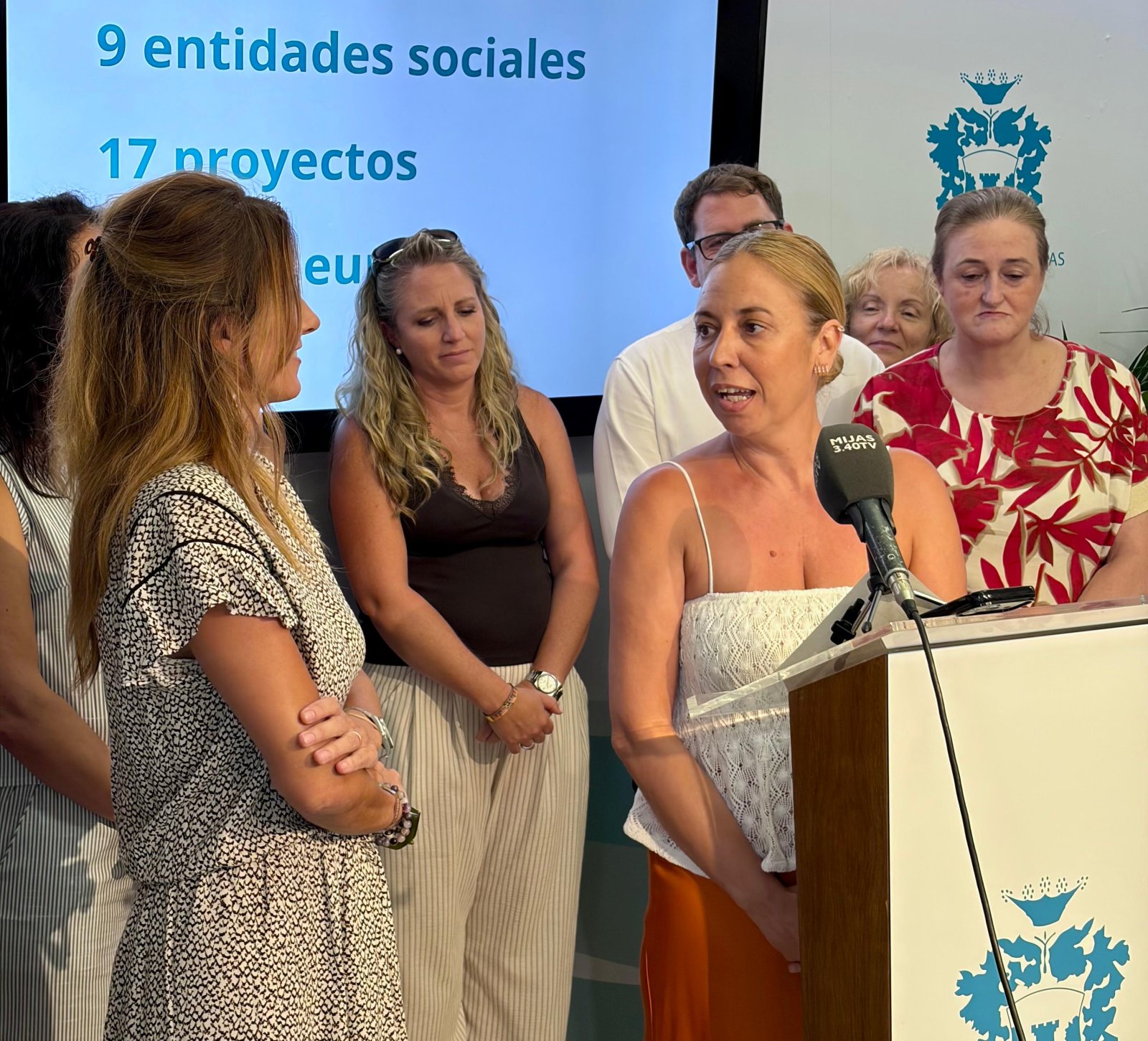 Una mujer habla ante un micrófono en un atril, rodeada de varias personas, con una pantalla detrás que muestra texto sobre entidades y proyectos.