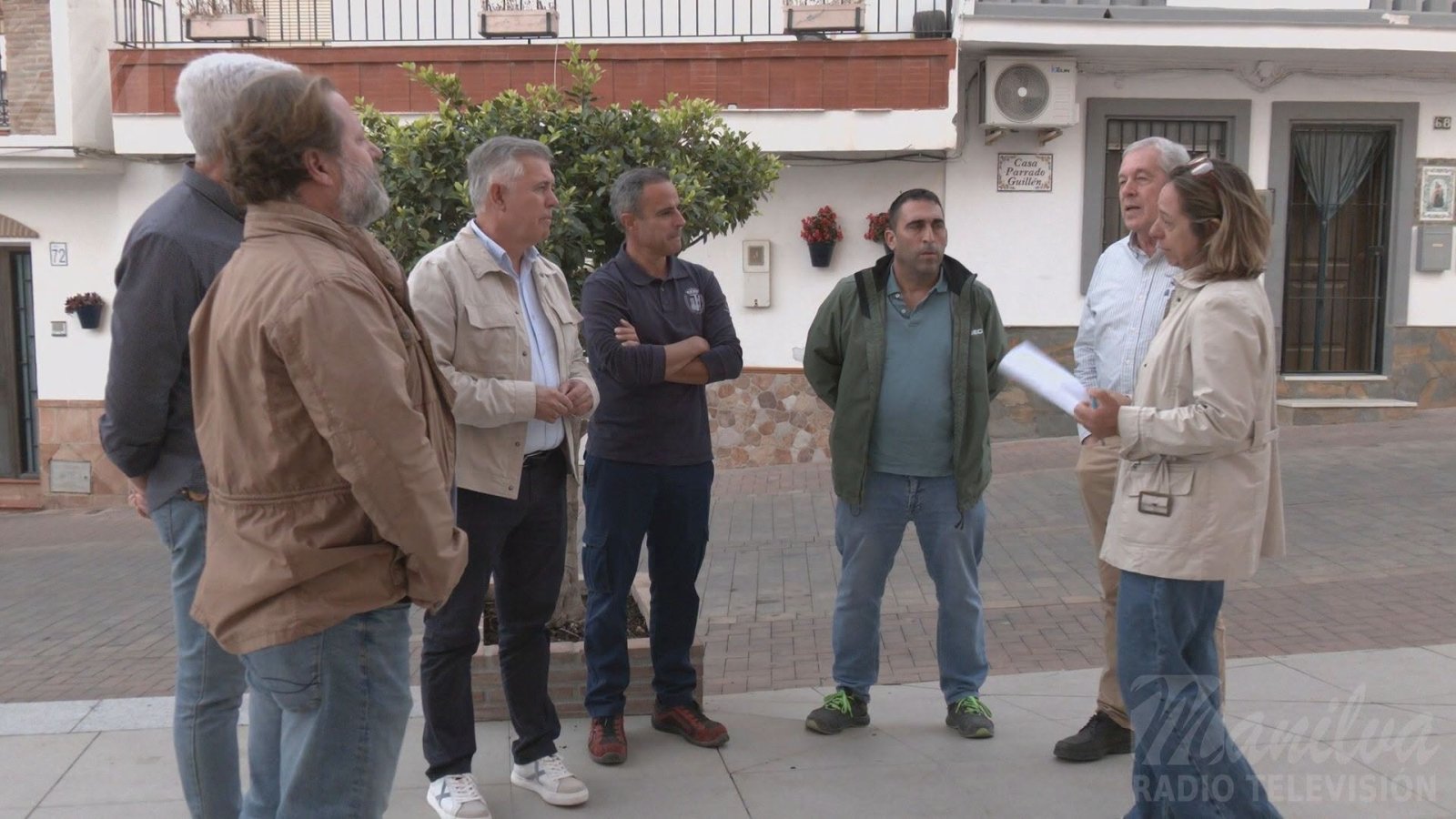 Seis hombres y una mujer de pie conversando en una calle empedrada, con fachadas blancas y un árbol al fondo.