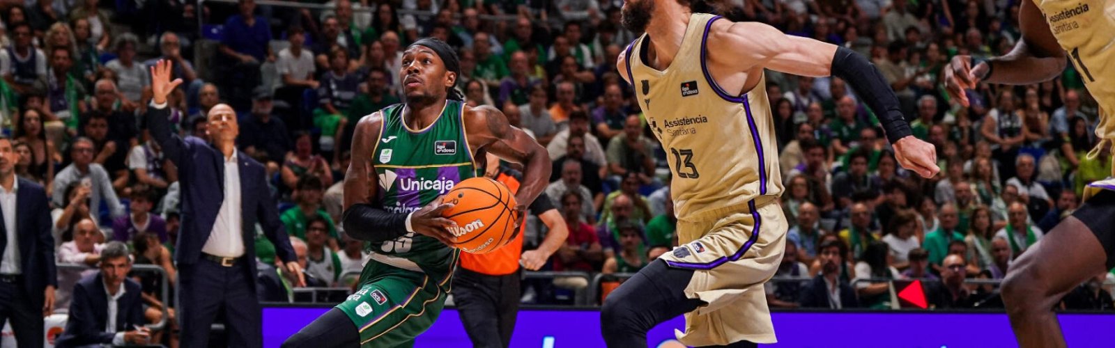 Un jugador de baloncesto con camiseta verde y balón en las manos ante un defensor de camiseta dorada, con público al fondo.