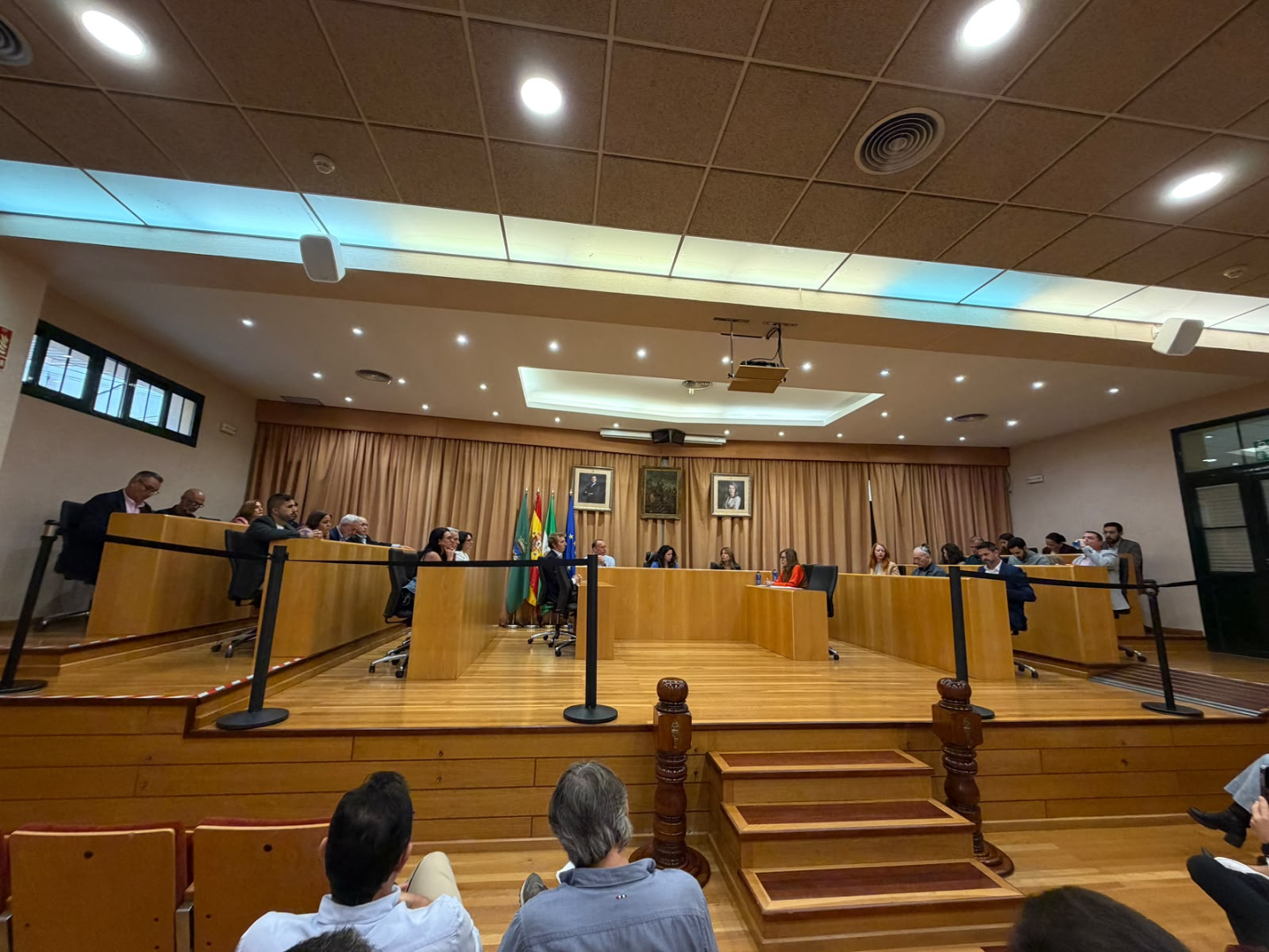 Sala de plenos con varias personas sentadas tras una bancada de madera en forma de U, banderas y retratos al fondo, y público en primer plano.