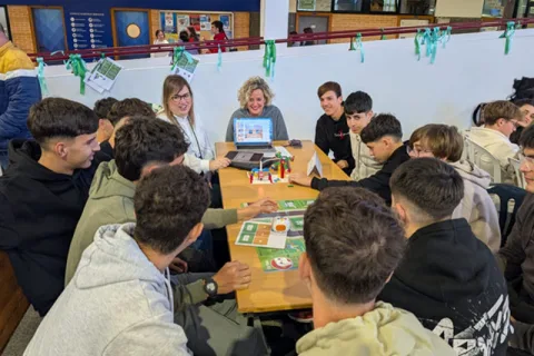 Jóvenes sentados alrededor de una mesa con un portátil y tableros de juego, junto a dos mujeres adultas, en un aula.