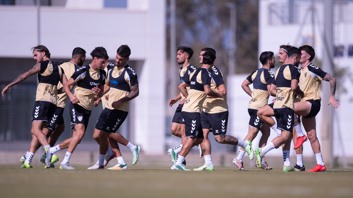 Varios futbolistas con camiseta beige y pantalón negro entrenan corriendo sobre el césped de un campo.