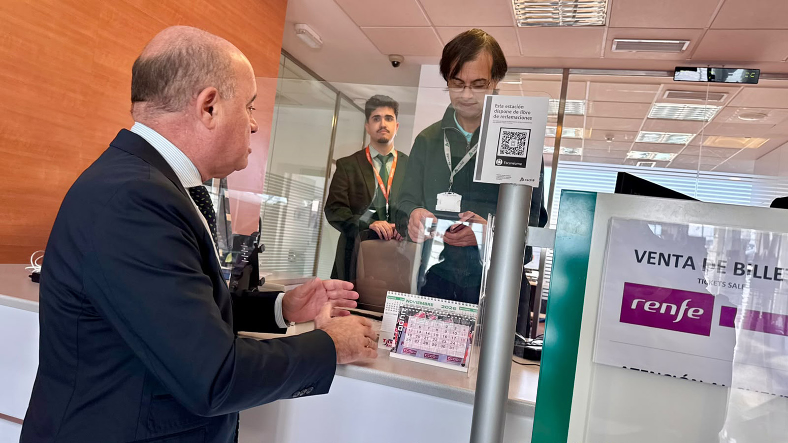 Un hombre trajeado habla en el mostrador de venta de billetes de Renfe con dos empleados tras la mampara.