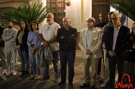 Un grupo de personas de pie y en silencio en una calle empedrada junto a una fachada encalada y plantas.