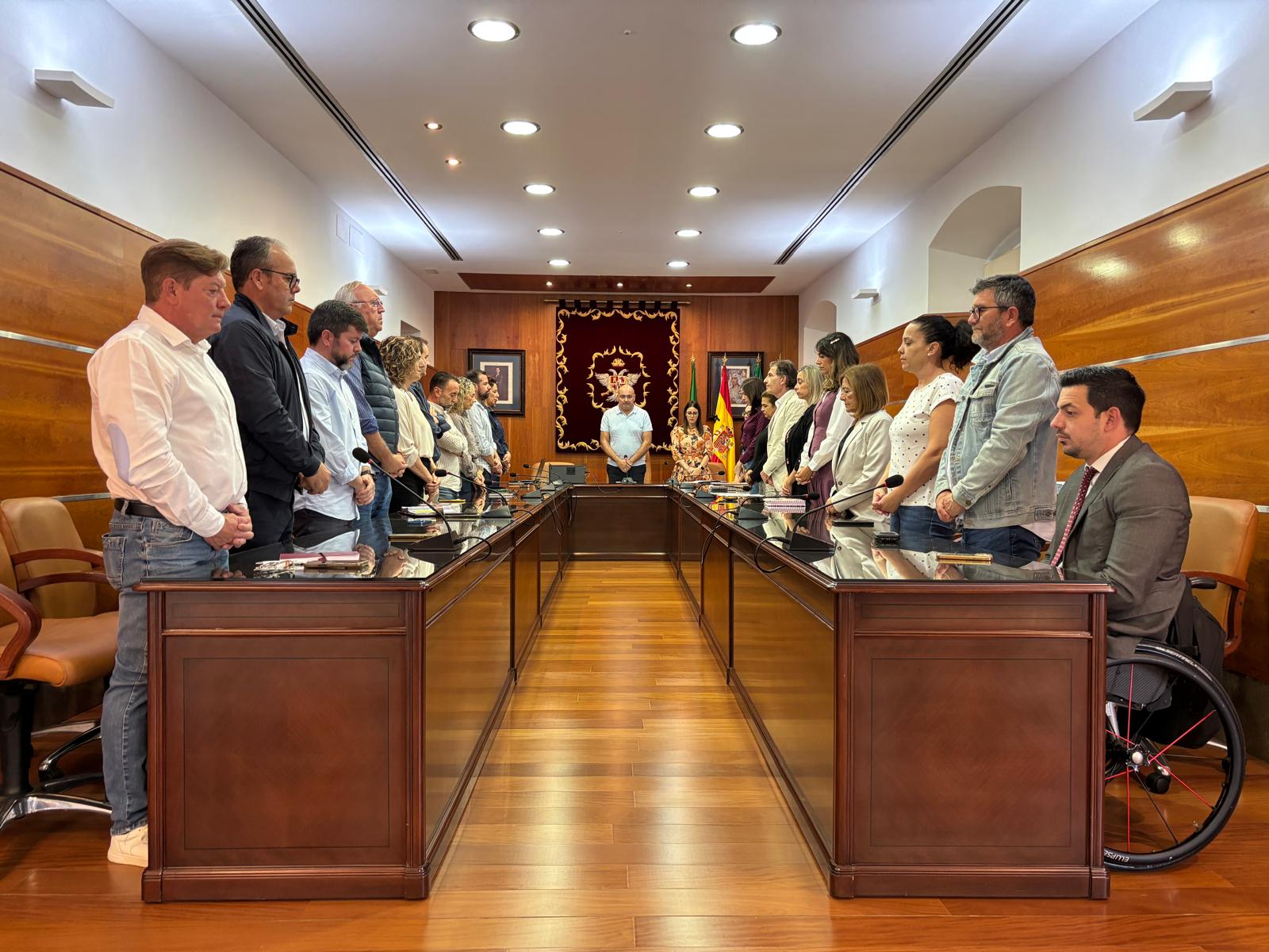 Un grupo de personas de pie junto a una mesa alargada de madera en una sala con banderas y un tapiz al fondo.