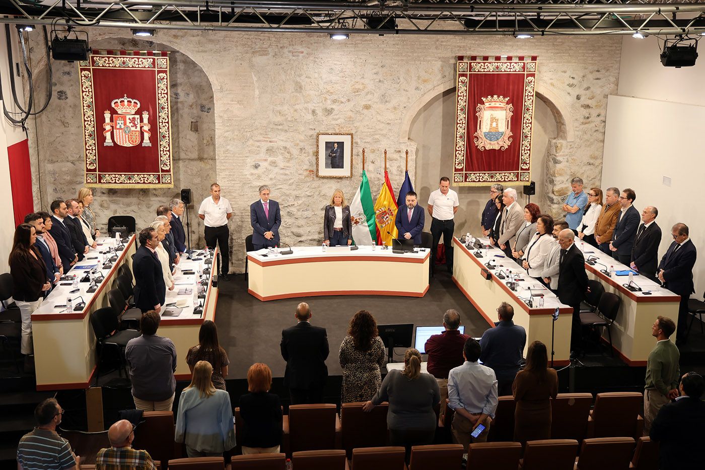 Personas de pie en un salón con muros de piedra, banderas de España, Andalucía y Europa, y dos tapices con escudos.