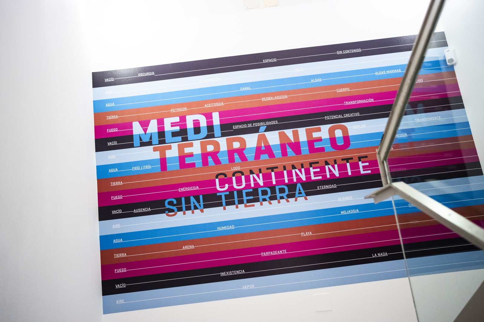 Cartel de pared con franjas horizontales de colores y el texto grande MEDITERRÁNEO CONTINENTE SIN TIERRA, con palabras sueltas en las franjas.