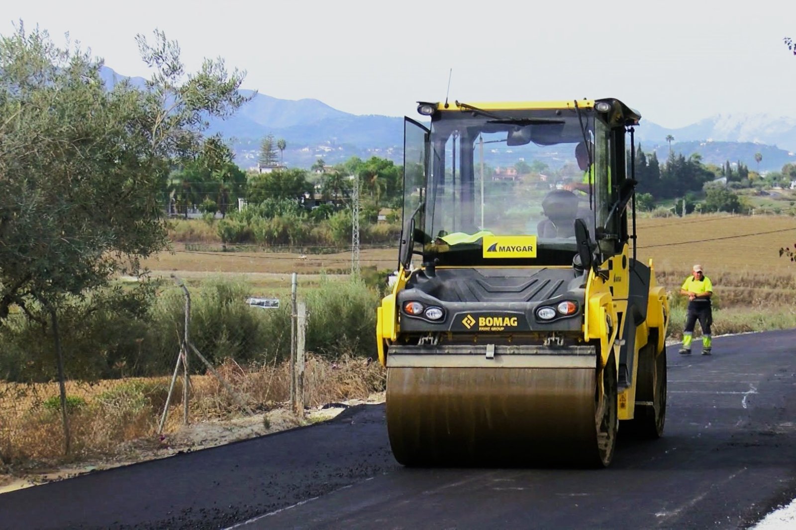 Un rodillo compactador amarillo sobre asfalto negro recién extendido, con un operario de chaleco reflectante al lado y campo y montañas al fondo.
