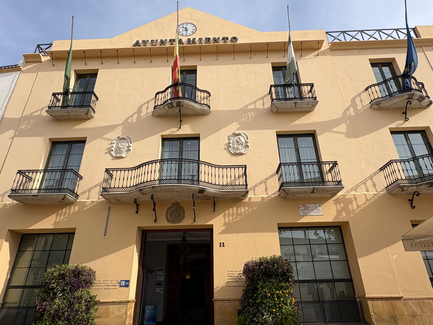 Fachada amarilla de un ayuntamiento con reloj, balcones de hierro, escudos y varias banderas en astas.
