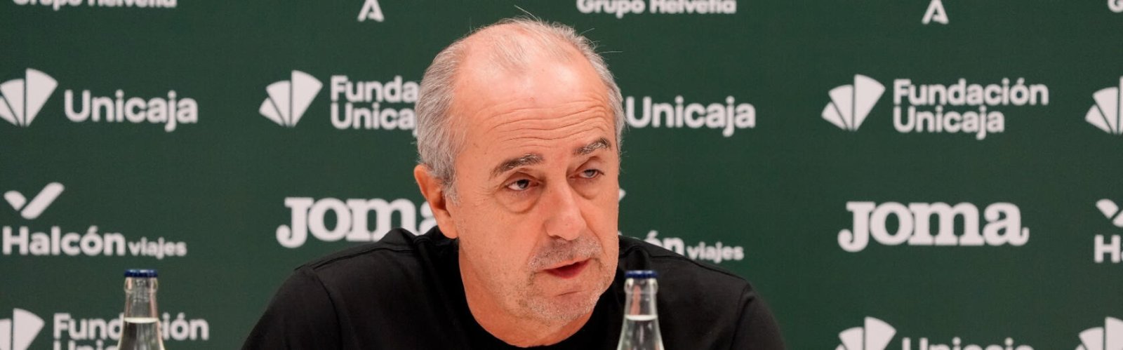 Hombre calvo con barba incipiente y camiseta negra hablando ante un fondo verde con logotipos de Unicaja, Joma y Halcón Viajes.
