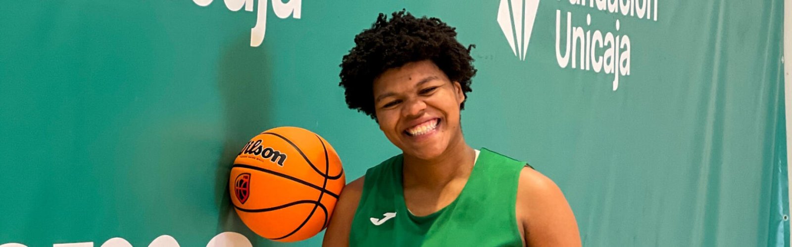 Una jugadora de baloncesto sonriente con camiseta verde sostiene un balón Wilson ante un fondo verde con el logotipo de Unicaja.